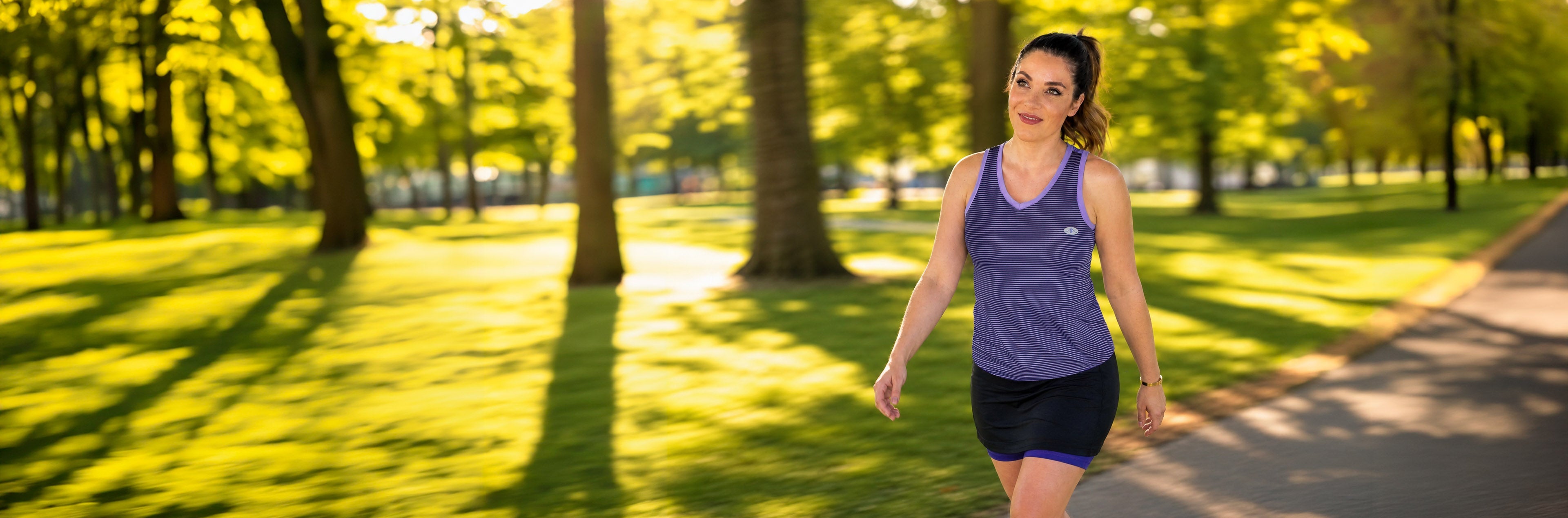 Woman walking on path in park in purple and lilac Alex + Abby active tank top and black and purple Pursuit Skort.
