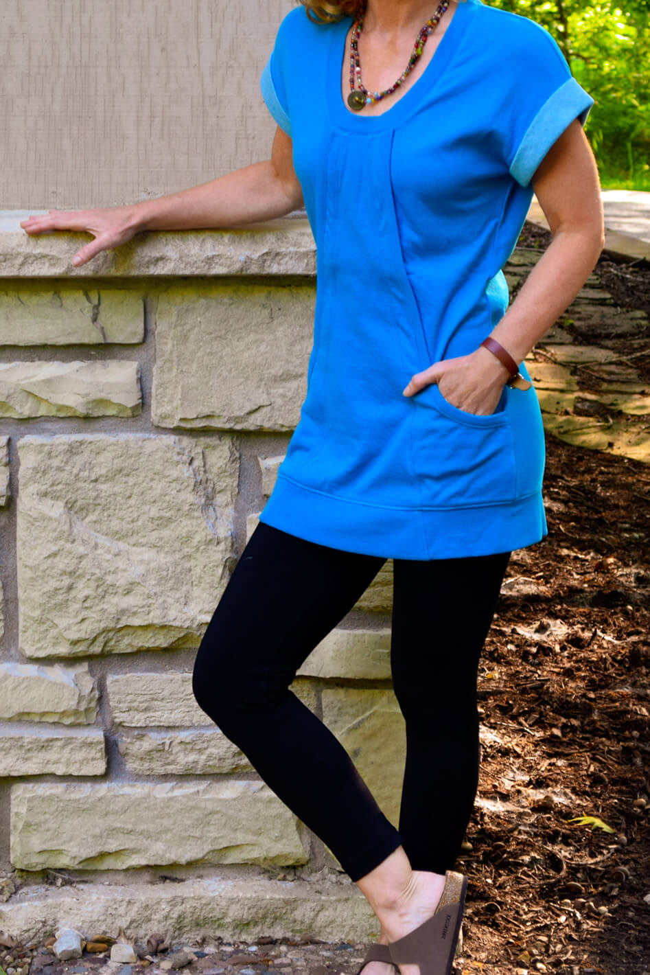 #color_malibu-teal A woman standing with one hand against a stone wall, wearing a short sleeve blue scoop neck tunic with pockets and a flat lay neckline.